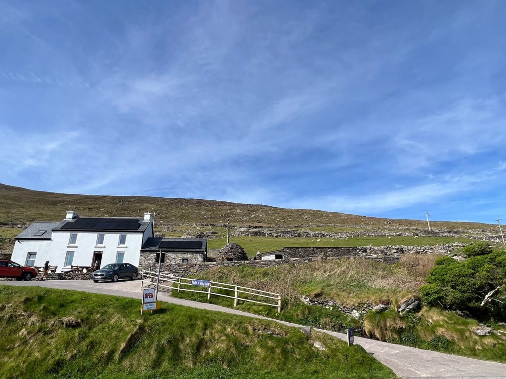 Rural landscape with a white house, open sign, car, and stone walls under a blue sky.